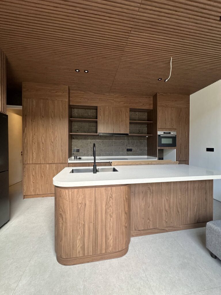 A beautifully designed kitchen in the Umalas Creek Villa Resort Custom Cabinet project, featuring a large curved island with a white quartz countertop, surrounded by warm walnut veneer floor-to-ceiling cabinets and a matching slatted wood ceiling. The kitchen showcases modern tropical luxury with integrated appliances and a stylish black faucet.