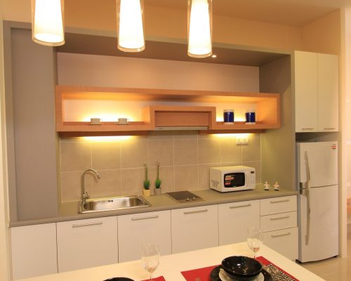 833 units KualaLumpur Hotel Fit Out 3 A modern compact kitchen and dining area featuring white lower cabinets, a light wood open shelf with integrated lighting, a stainless steel sink, and three contemporary cylindrical pendant lights over a white dining counter set for two.