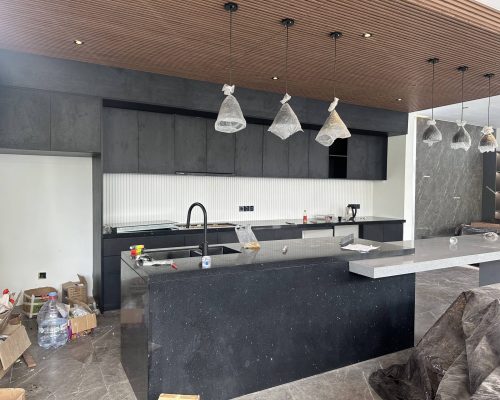 11 Sets Umalas Creek Villa Resort Custom Cabinet 4 A contemporary kitchen or bar area within the Umalas Creek Villa Resort Custom Cabinet project, featuring sleek matte dark grey cabinetry, a black speckled island countertop, and a matching slatted wood ceiling. Exposed pendant light fixtures are wrapped for protection, indicating ongoing construction, while the space hints at sophisticated, modern design.