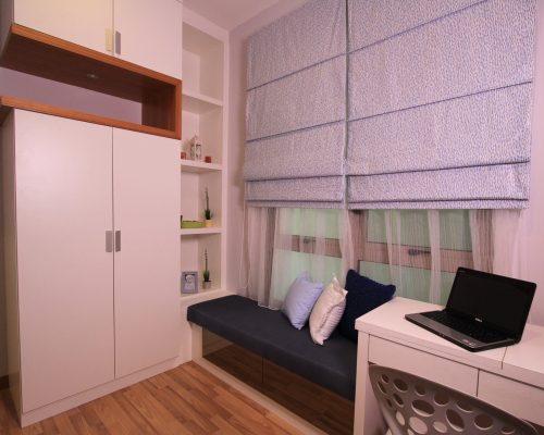 833 units KualaLumpur Hotel Fit Out 7 A bedroom corner with a large white built-in wardrobe and open shelving, next to a window with a blue patterned Roman shade, and a dark upholstered window bench with cushions and a laptop on a white desk extension in KualaLumpur hotel fit out.